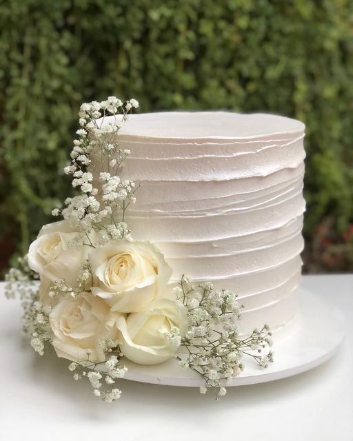 Elegant white textured wedding cake decorated with white roses and baby's breath on a simple white stand.