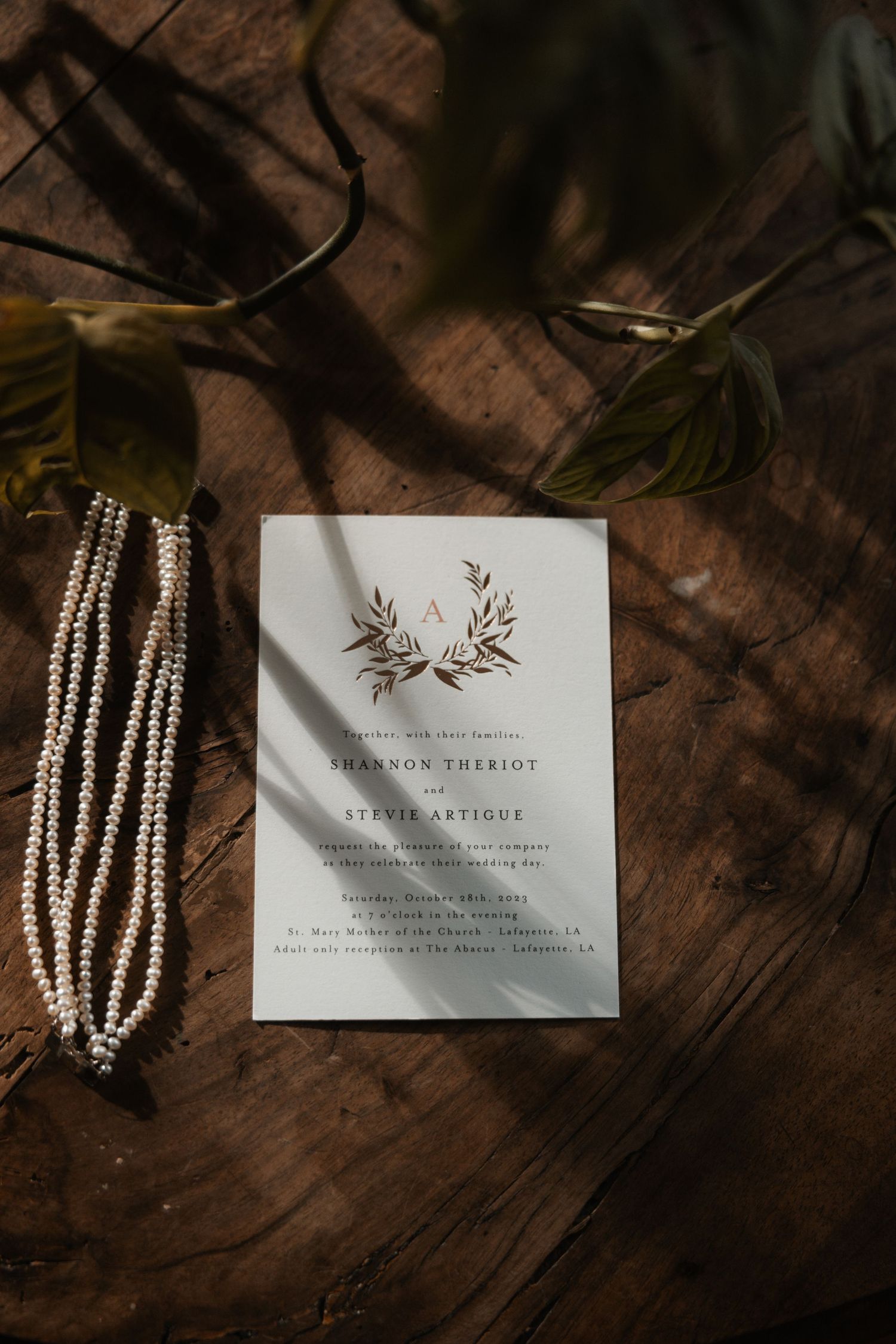 Pearl necklace and elegant wedding invitation with leaf design on rustic wooden background in moody lighting.