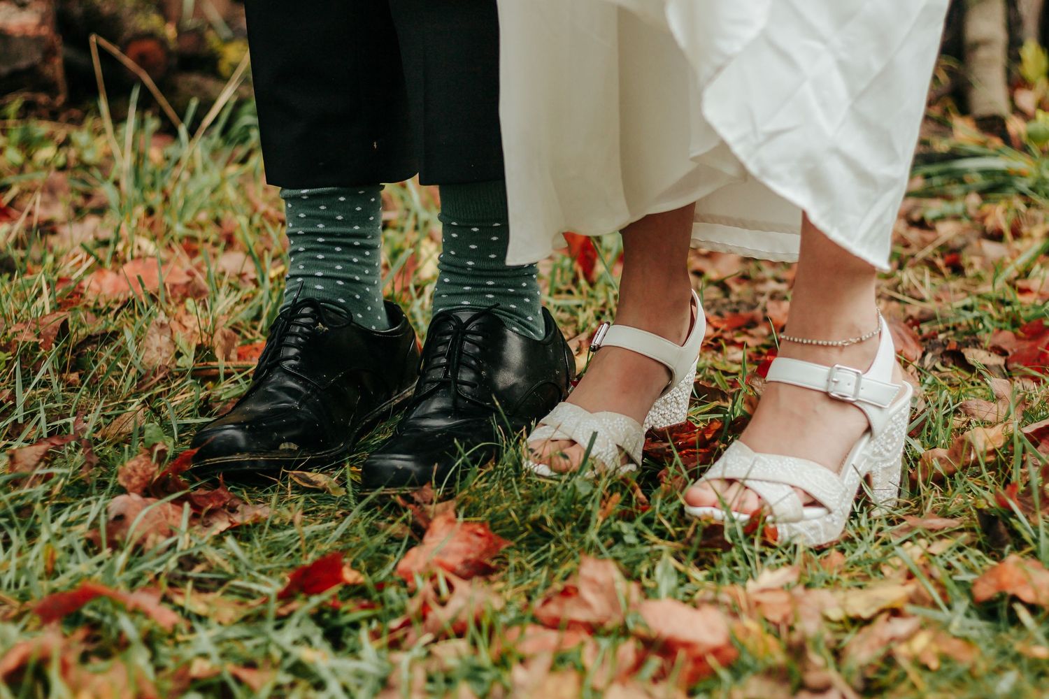Close-up of contrasting wedding footwear showing black boots and white sandals on fallen autumn leaves.