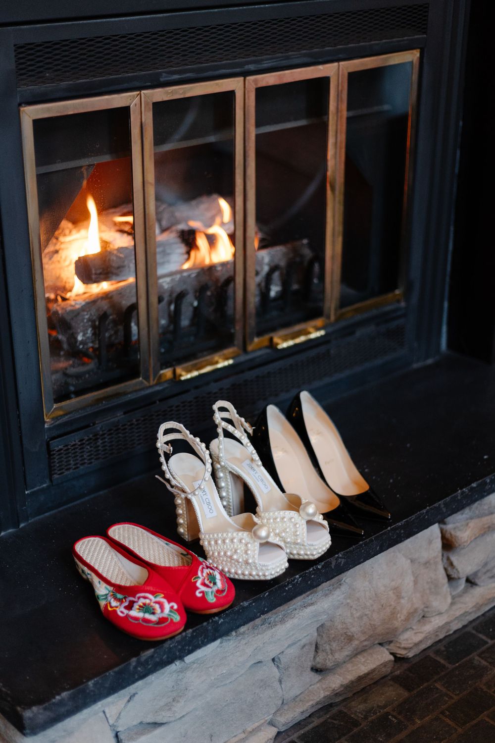 Three pairs of shoes arranged near a glowing fireplace, including red flats and white heels.