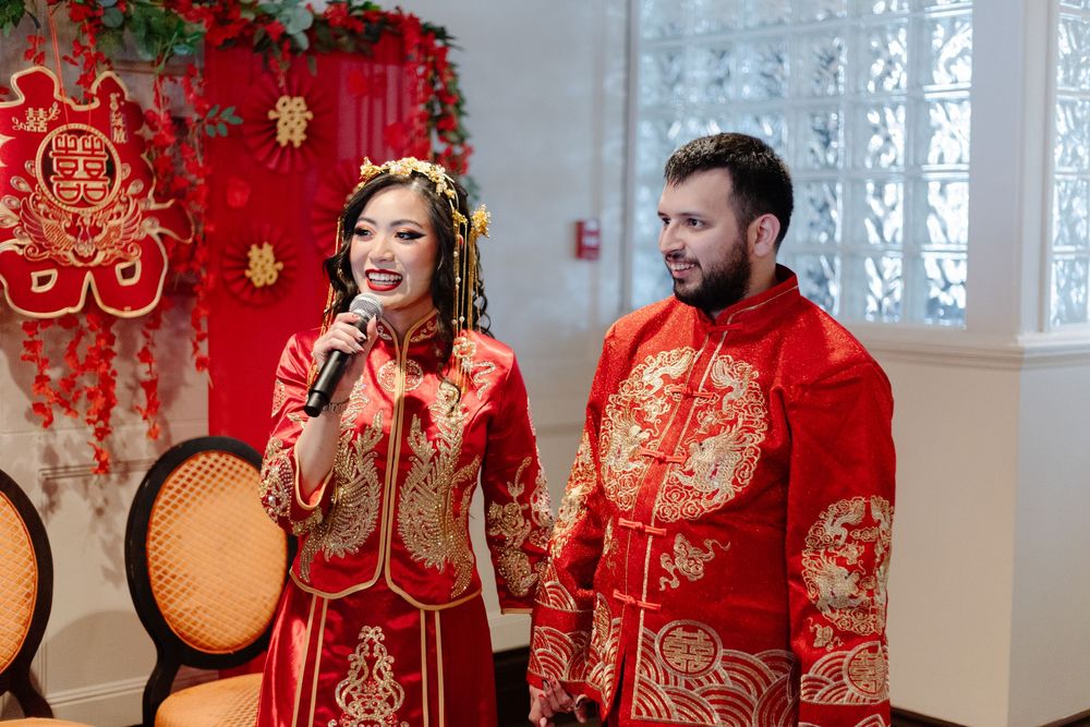 Couple in traditional red Chinese wedding attire with gold embroidery smiling during ceremony.