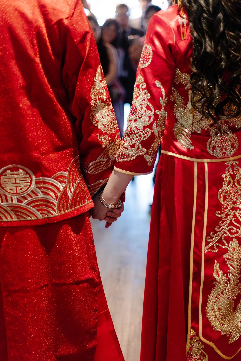 Couple in traditional red Chinese wedding attire with gold embroidery holding hands during ceremony.