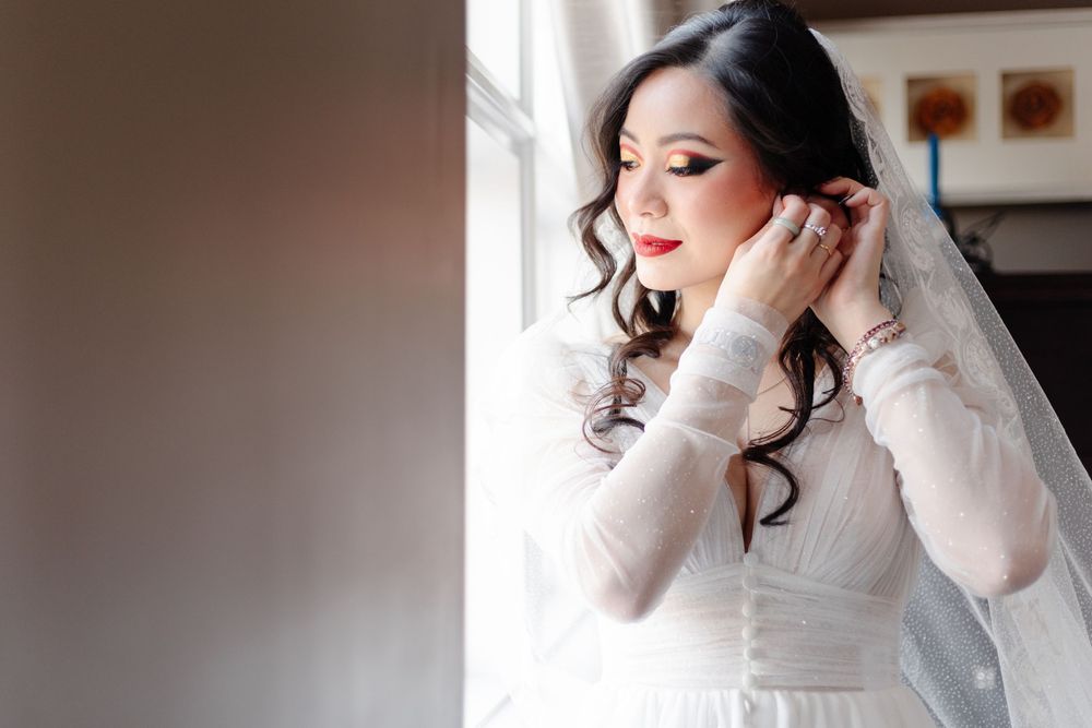 Beautiful Asian bride in white lace gown adjusting earrings by window on wedding day.