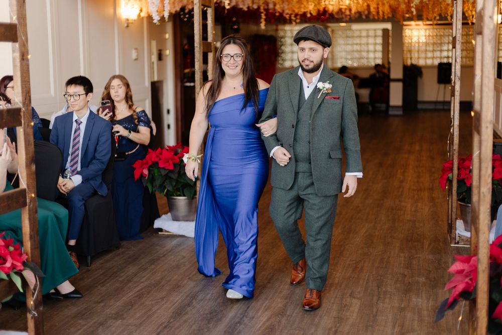 A couple walks arm-in-arm down a decorated wedding venue aisle, woman in blue gown, man in green suit.