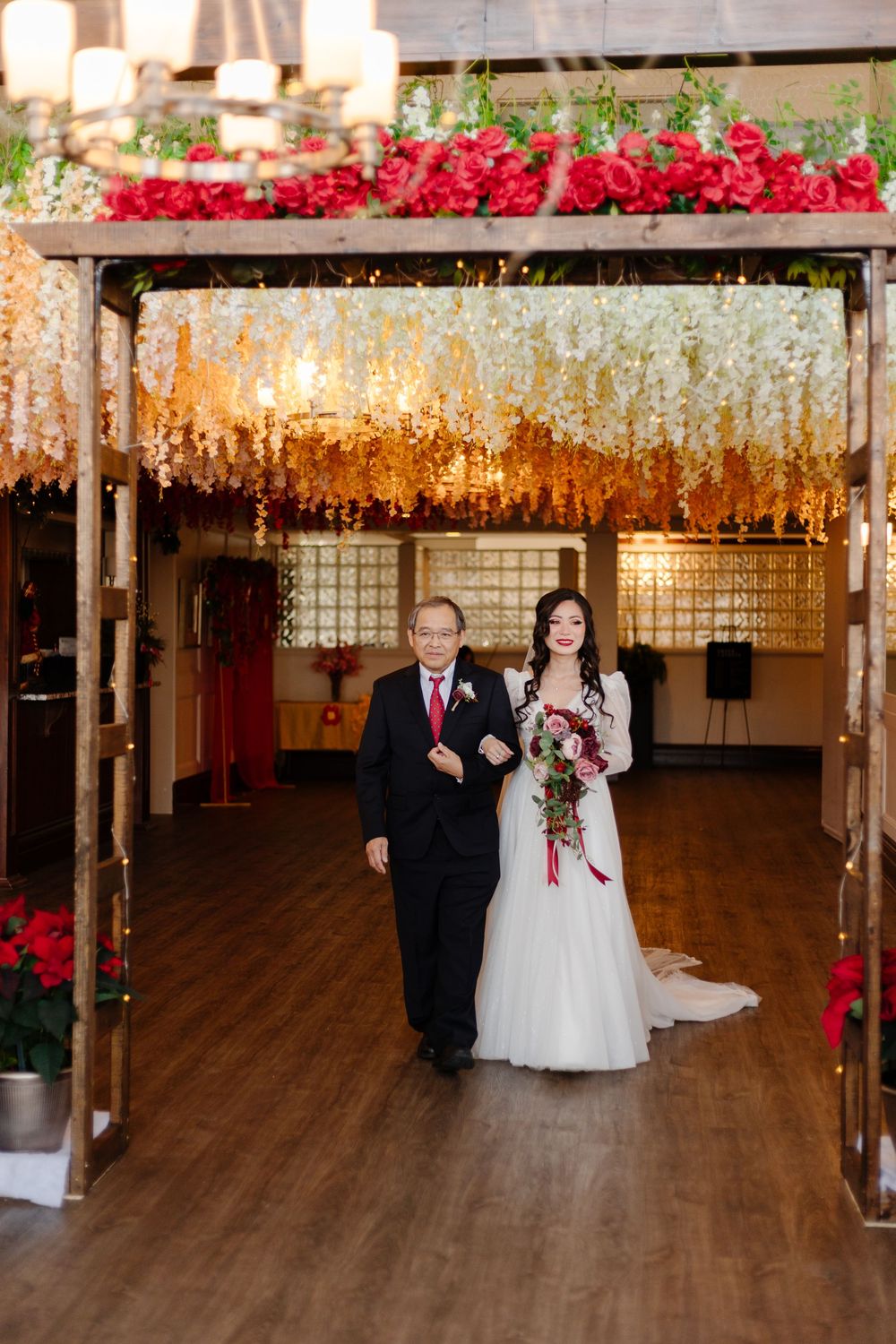 Bride in white gown walks down aisle with father under floral arch with red roses.