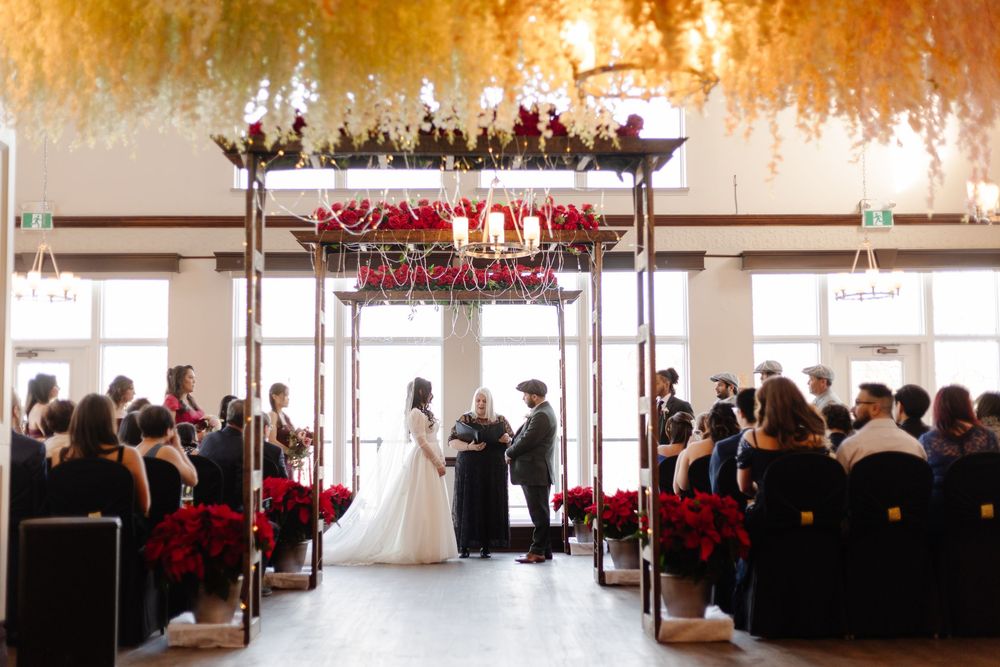 Elegant wedding ceremony with red floral arrangements, hanging florals, and guests seated in a decorated venue.