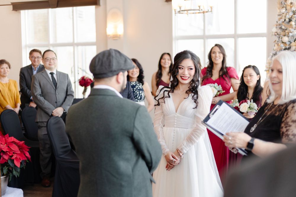 Bride smiling at groom during indoor winter wedding ceremony with guests and bridesmaids in red dresses.