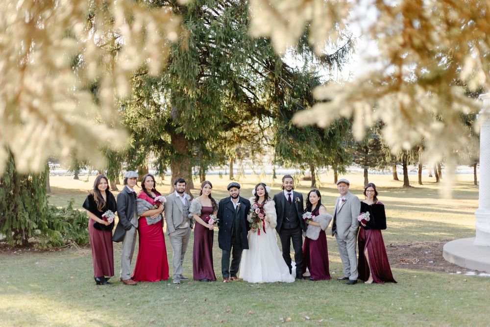 Wedding party in burgundy and gray attire posing outdoors under autumn tree branches.
