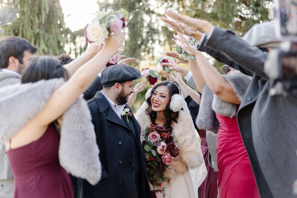 Newlyweds walk through cheering guests outdoors in winter attire, bride holding a bouquet.