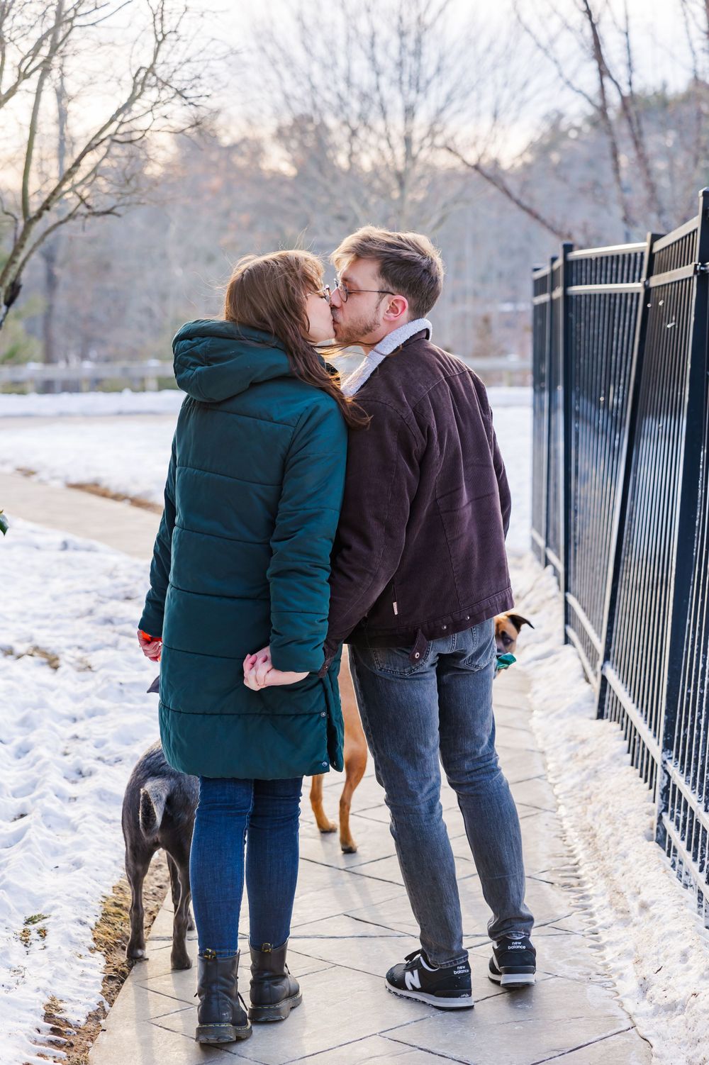 A couple shares a winter kiss while walking their dog along a snowy path with iron fence.