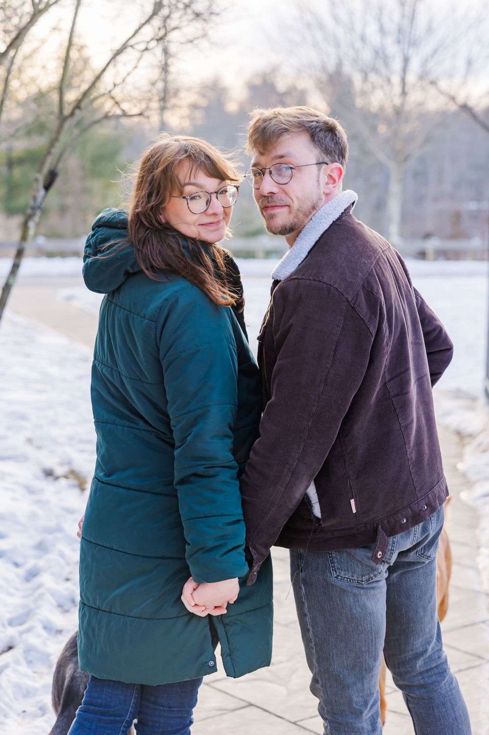 A couple wearing winter coats stands together in a snowy outdoor setting while looking at the camera.