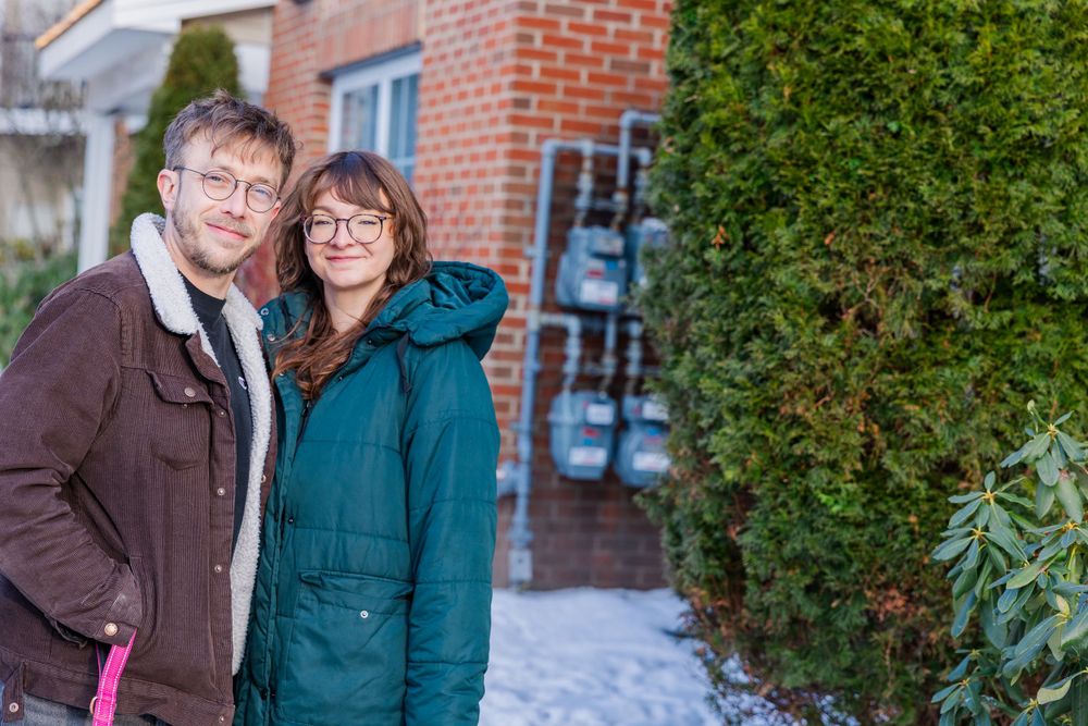 A couple poses outside a brick building wearing winter coats.