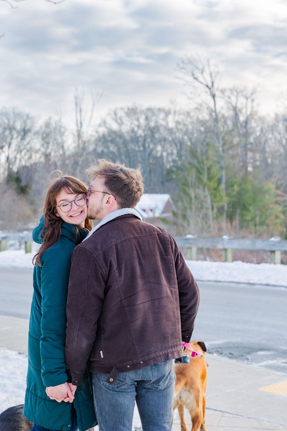 A couple embraces outdoors in winter coats with a dog visible in the background.