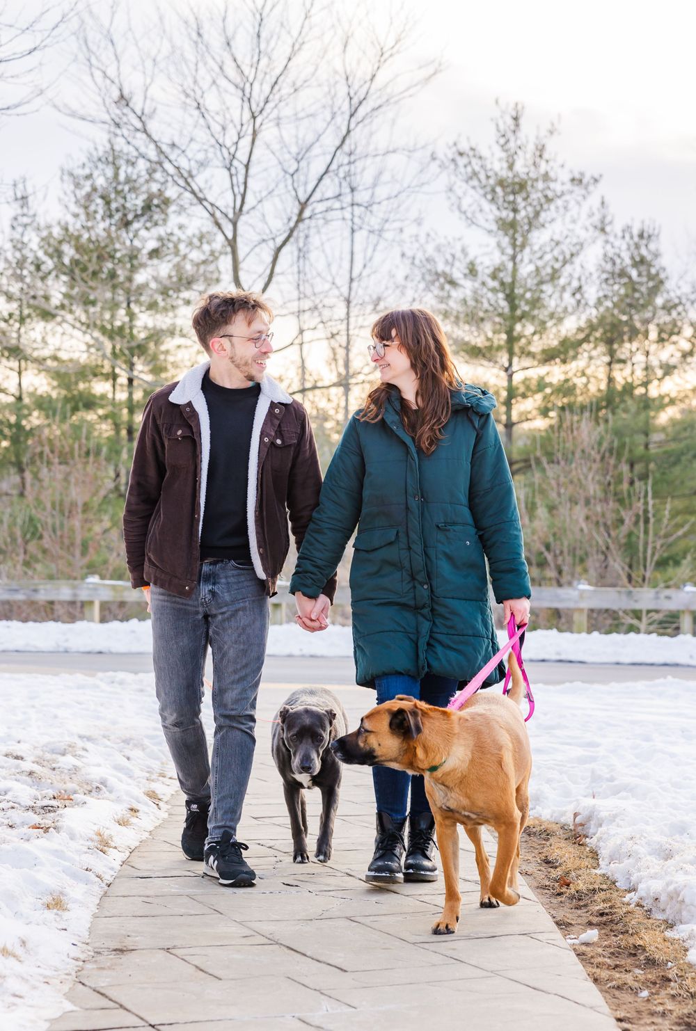 A couple walks their two dogs on a snowy path during sunset.