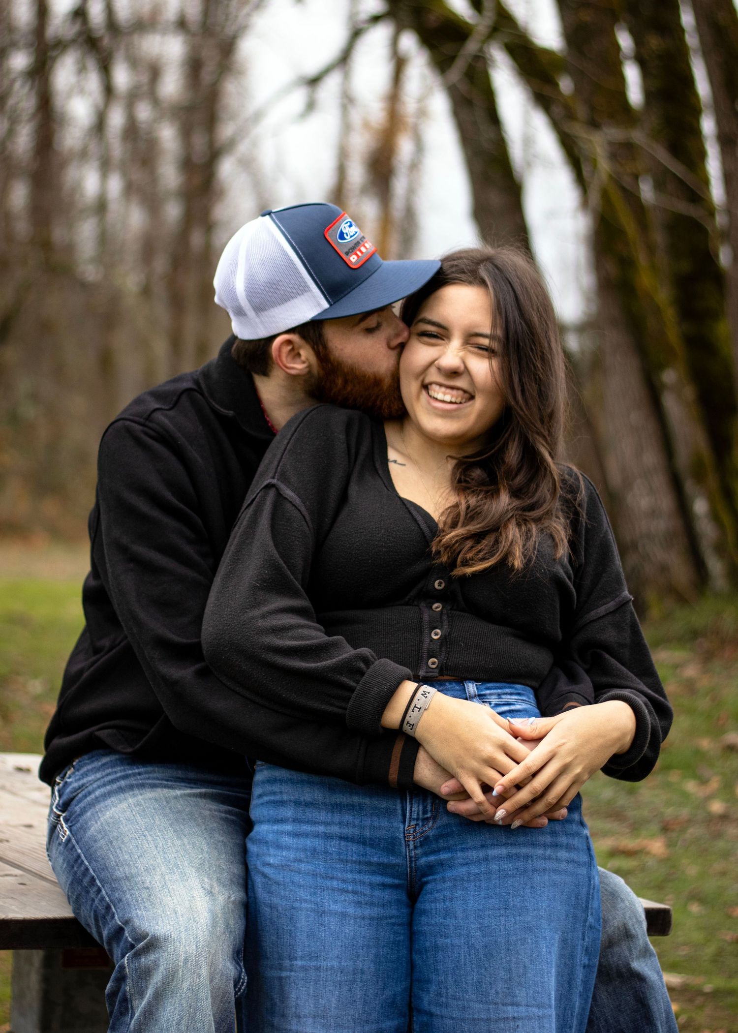 a man hugs a woman from behind and kisses her cheek as she smiles. they are dressed in black shirts and blue jean and are by trees
