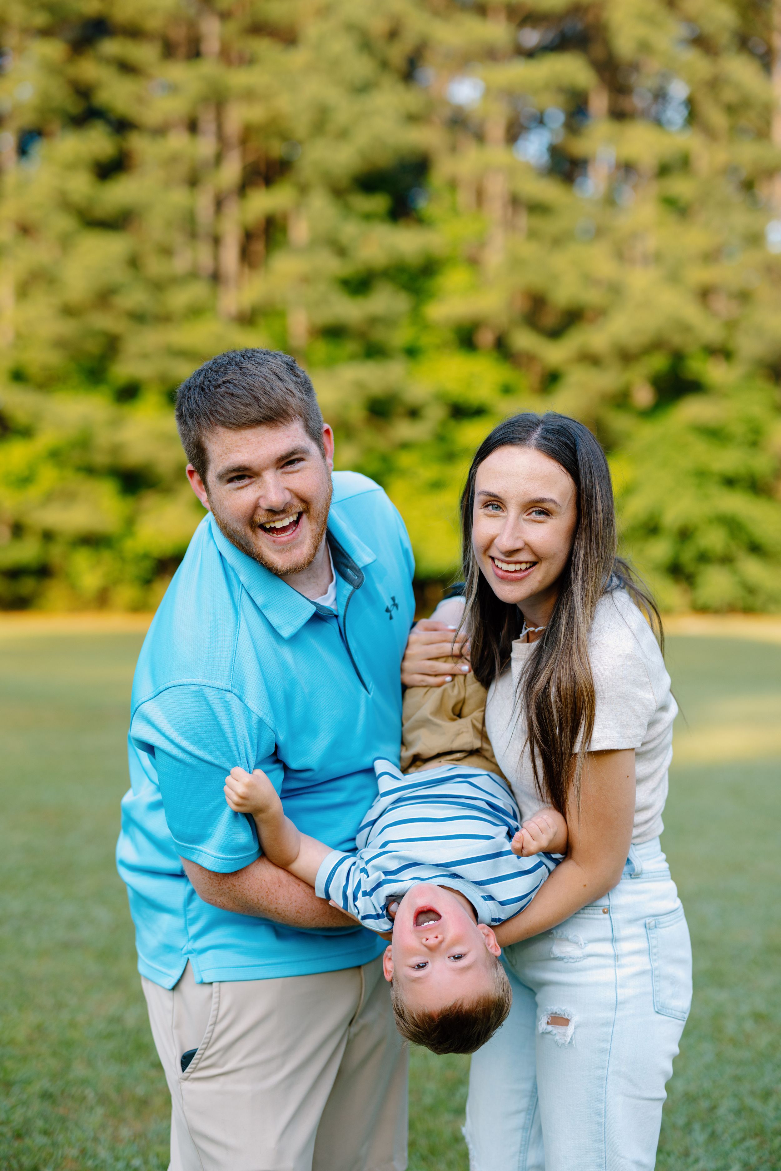 Playful family moment outdoors with child held upside down against green forest background.