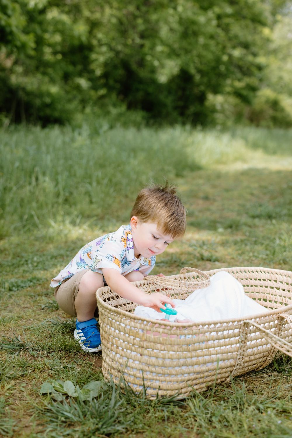 Little one in floral shirt crouches by large woven basket on grassy ground during summer day.
