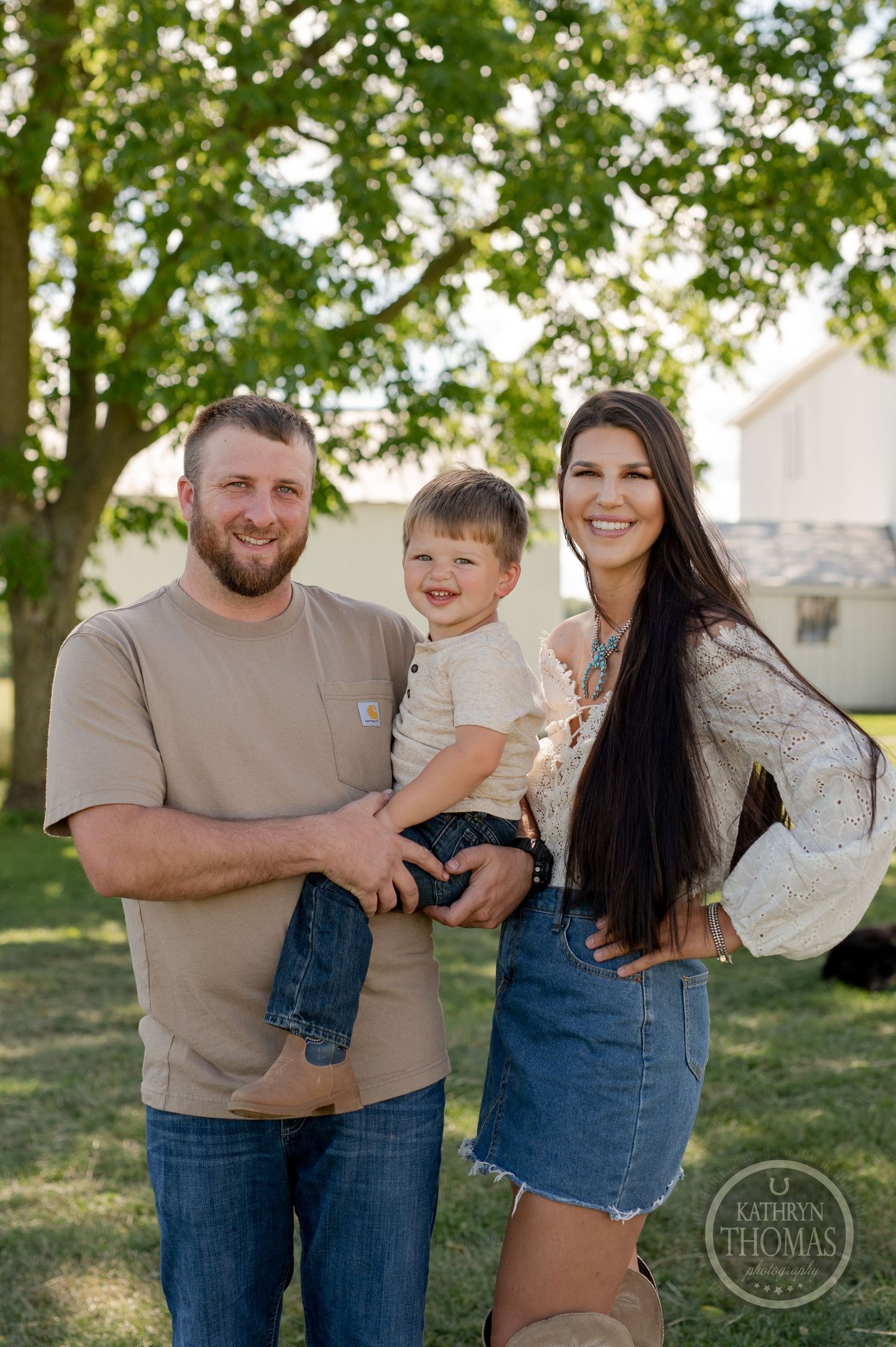 A family poses together on their farm property with trees and buildings behind them.