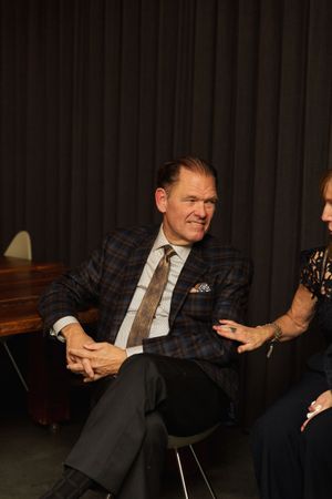 A person in a plaid suit jacket and tie sits relaxed in conversation against a dark backdrop.