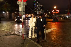 Couple in formal attire standing on wet city street at night with illuminated buildings.