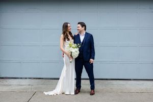 Bride in white gown and groom in navy suit share intimate moment against grey wall.