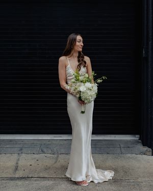 Bride in form-fitting white gown carries white floral bouquet against dark background.