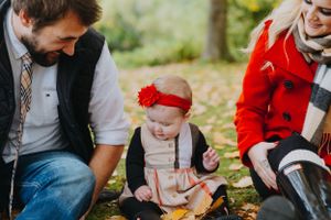 A small toddler wearing a red headband explores fall leaves outdoors with adults during autumn.