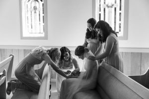 A heartwarming moment shows wedding party members helping with dress preparations in a church room.