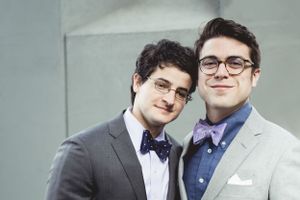 Two people in formal suits and bow ties share an intimate moment against a grey background.