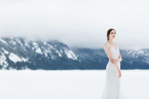 Bride in white dress stands against snowy mountain landscape.