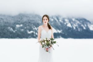 Bride in white dress holds bouquet against snowy mountain landscape.