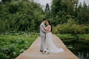 A couple in wedding attire embraces on a wooden dock surrounded by green nature.
