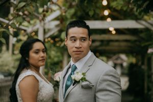 A couple in wedding attire poses together outdoors with string lights in the background.