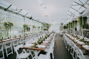 Elegant greenhouse wedding reception setup with long tables, white chairs and hanging greenery decorations.