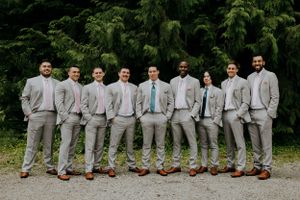 A group in matching light gray suits and brown shoes pose against evergreen trees.
