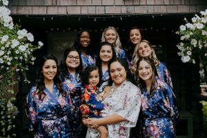 Group wearing floral blue robes gather around central figure in white on porch with flowers.