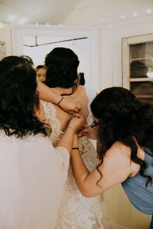 People helping with wedding dress preparations in an indoor setting with string lights.