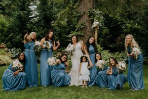 Wedding party poses playfully on grass with bridesmaids in blue dresses and white bouquets.