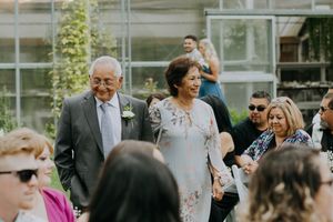 Wedding guests gather outdoors near a modern building with glass walls and climbing vines.
