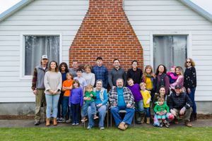Large multi-generational group poses together outside brick and white siding building.