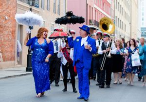 Festive parade performers in blue and sparkly outfits dancing with decorative parasols down a historic street.