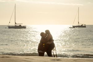 A silhouetted couple embraces on the beach at sunset with boats anchored in the bay.