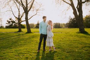 A romantic couple stands in golden sunlight on grassy hill with bare trees silhouetted behind them.