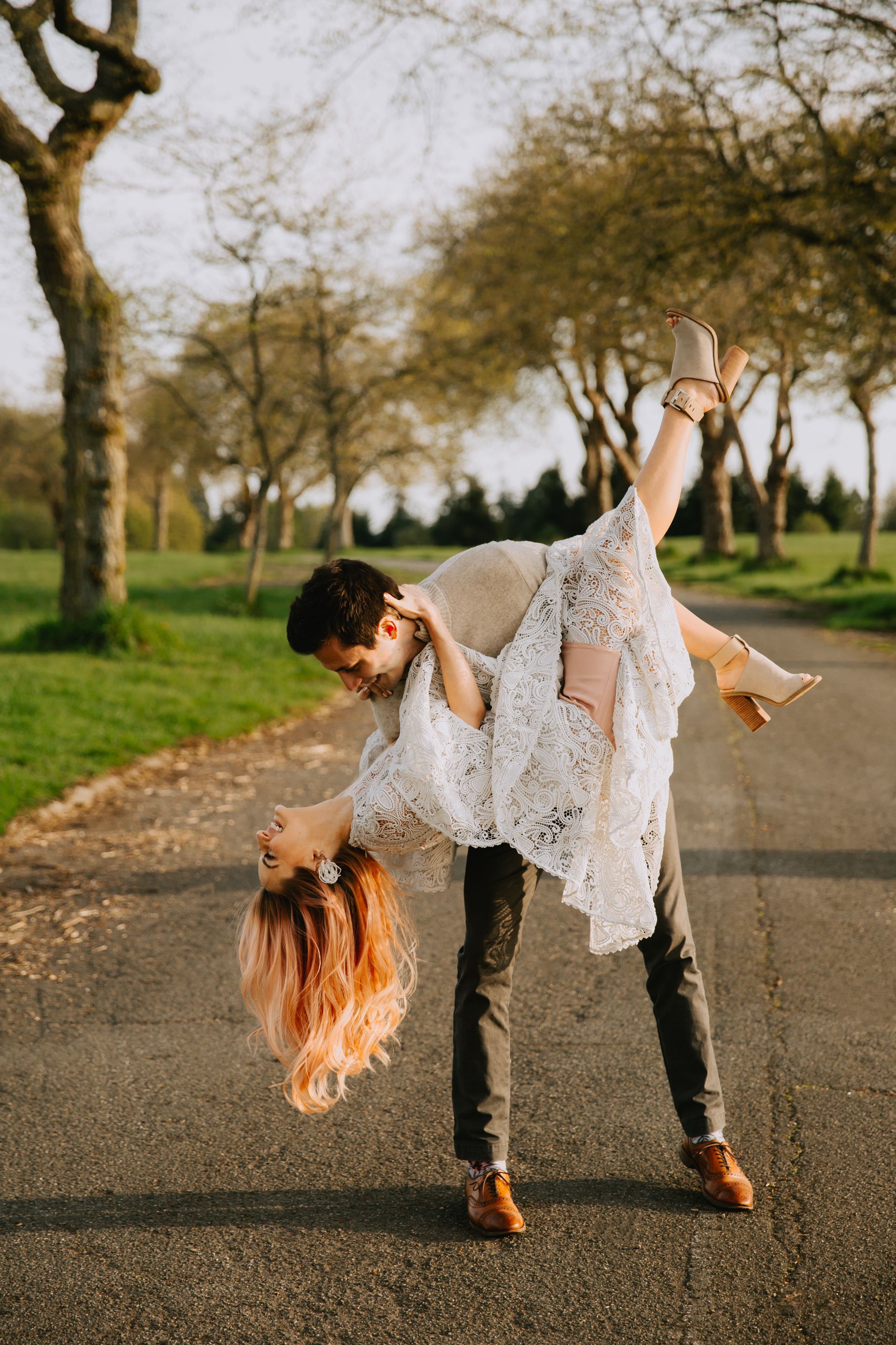 Playful couple performs dramatic dip dance move on tree-lined path at sunset.