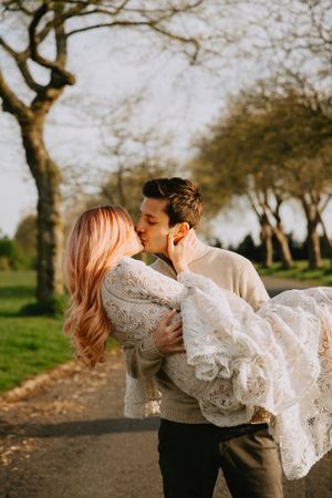 Romantic couple embraces in white lace dress and sweater on park path during sunset.