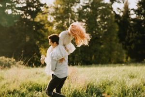 Joyful couple spins in golden meadow during sunset with evergreen trees in background.