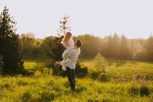 Couple twirls together in sunlit field with evergreen forest backdrop at golden hour.