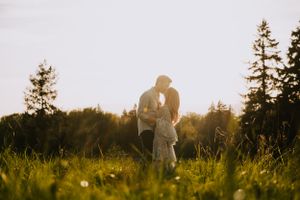 Silhouetted couple embraces in tall grass during golden hour with pine trees in background.
