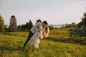 A couple embraces in a sunlit meadow with long grass and trees in the background during golden hour.