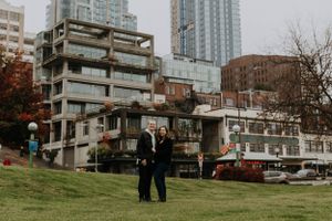 A couple standing on a grassy hill with urban architecture and high-rise buildings in the background.